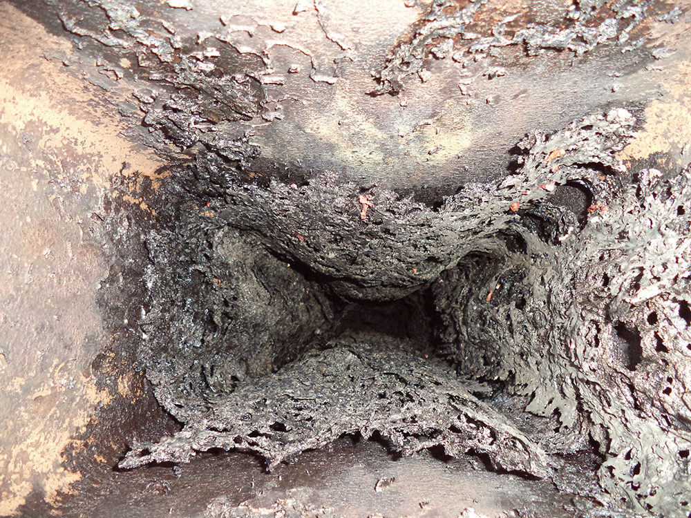 An interior view looking down a square metal flue heavily clogged with a thick, crusty, and porous buildup of black creosote, severely restricting the opening - juniatachimne1.wpenginepowered.com