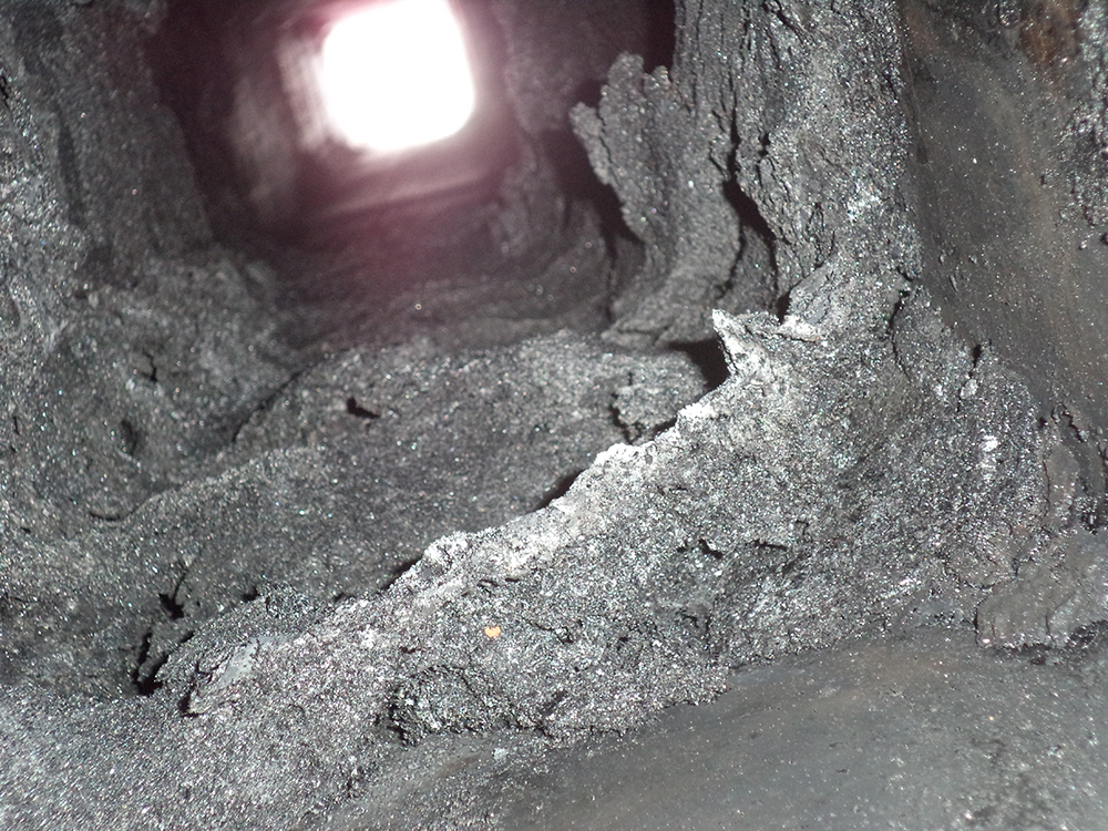 An interior view looking down a square metal flue heavily clogged with a thick, crusty, and porous buildup of black creosote, severely restricting the opening - juniatachimne1.wpenginepowered.com