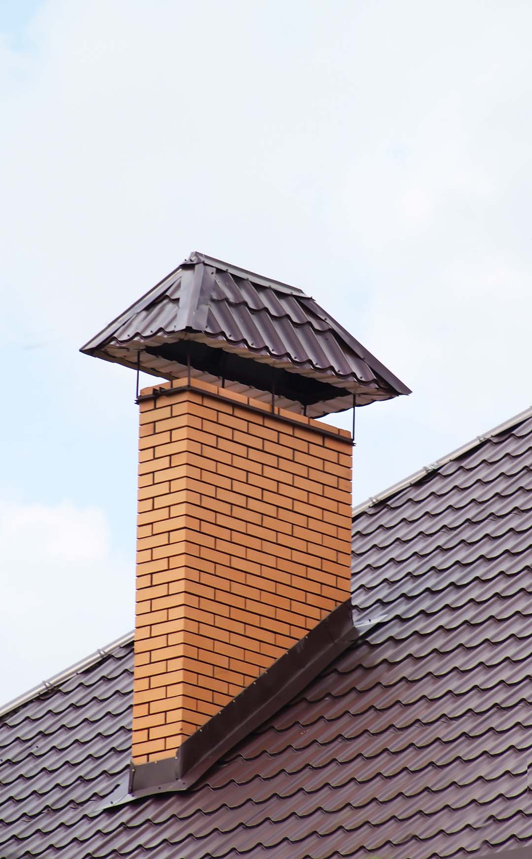 An orange brick chimney rises from a dark brown tiled metal roof, topped with a matching dark brown protective cap against a pale blue sky - juniatachimne1.wpenginepowered.com