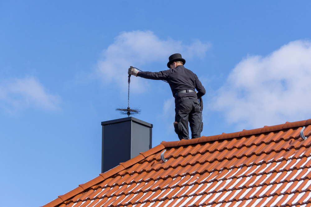 A chimney sweep wearing traditional black clothing and a top hat stands on a red-tiled roof, lowering a wire brush into a chimney against a bright blue sky - juniatachimne1.wpenginepowered.com
