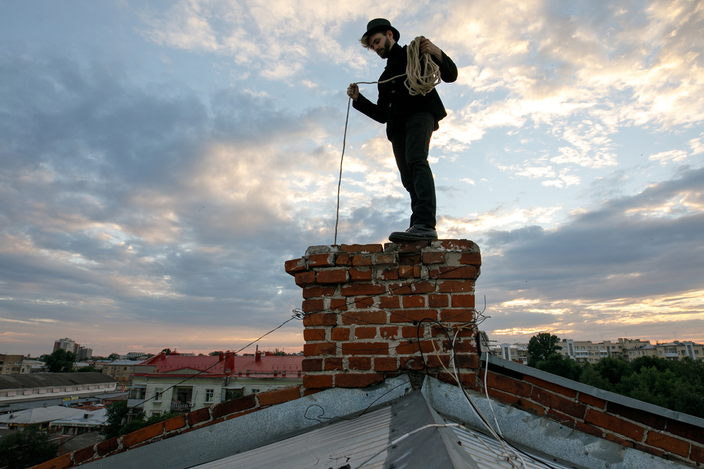 A man in a top hat and dark clothing stands atop a red brick chimney on a rooftop, lowering a rope down inside - juniatachimne1.wpenginepowered.com